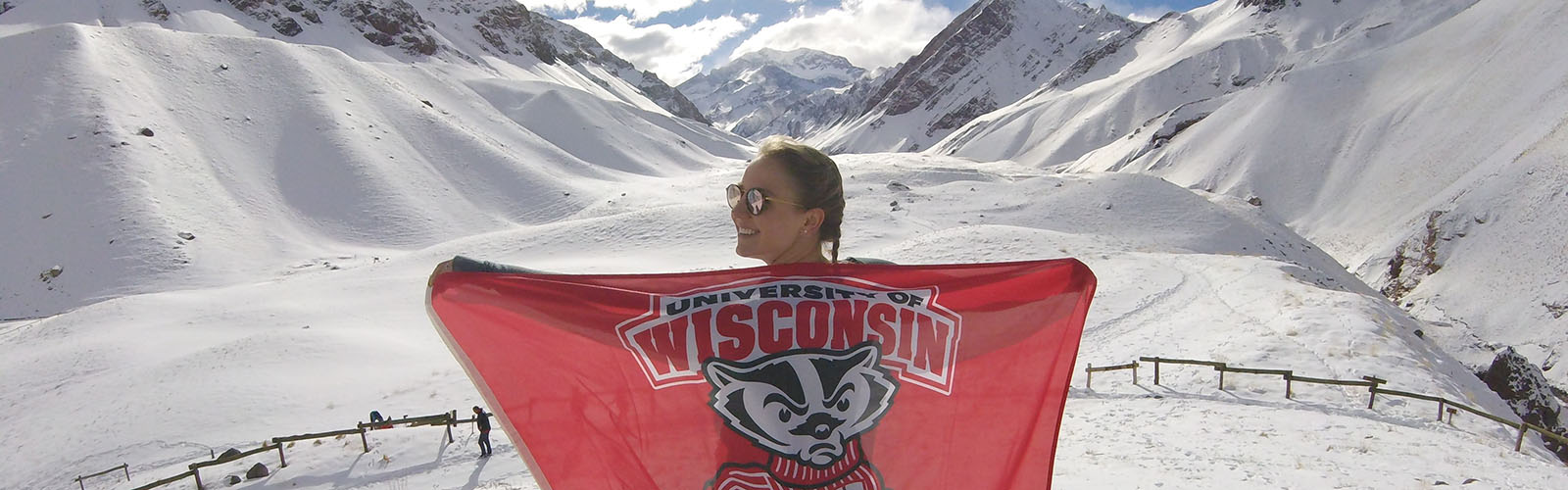 A student is holding a UW-Madison flag behind them and is looking over their shoulder. They are in front of a mountain range. 